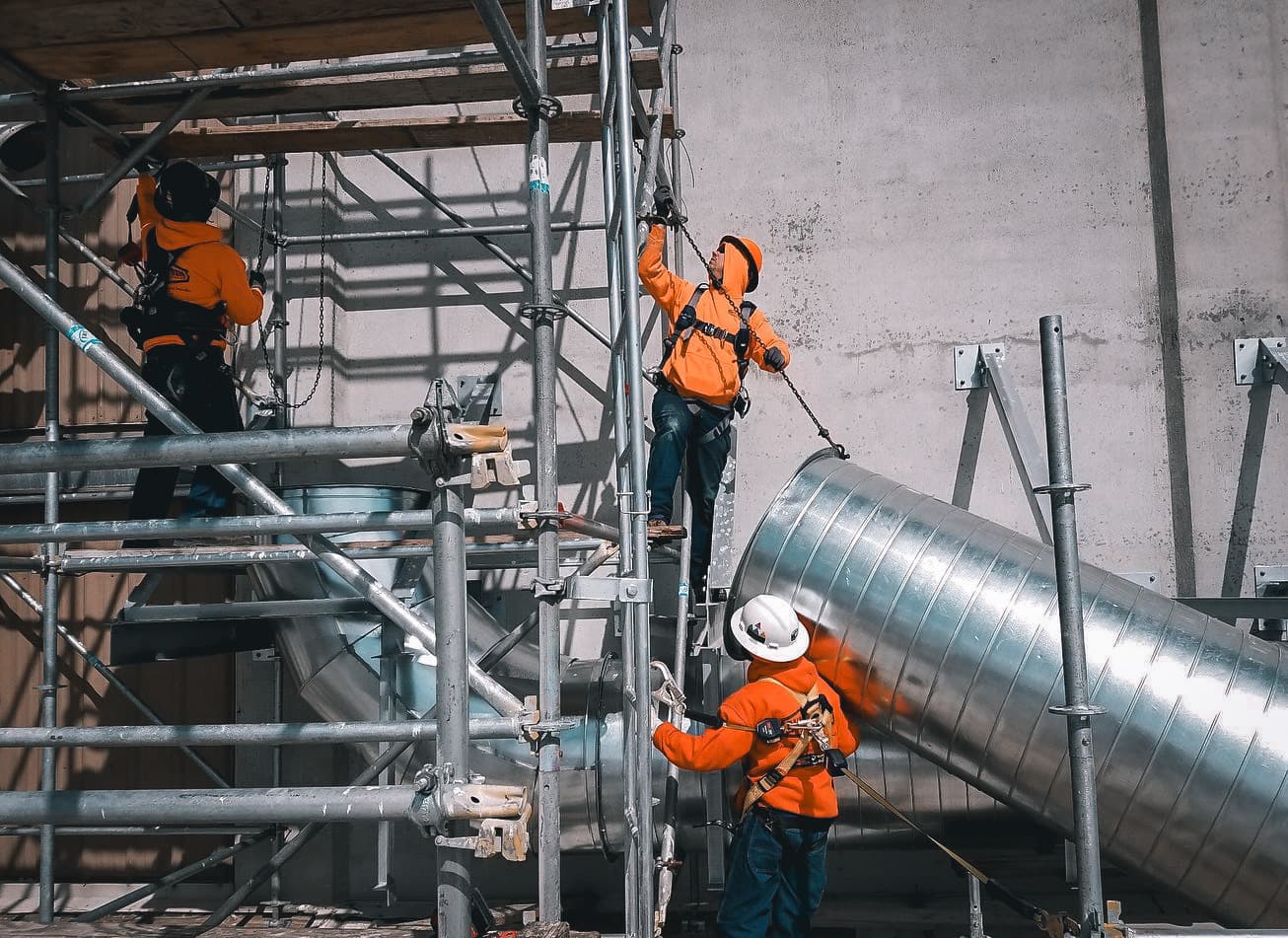 Three HVAC Technicians in orange safety gear installing metal ductwork and scaffolding inside an industrial building.