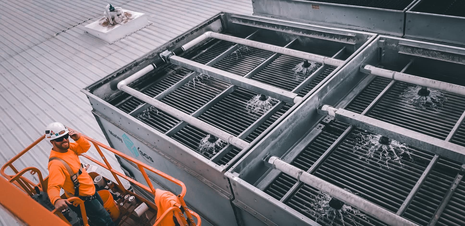 Worker in safety gear standing on lift inspecting active cooling tower system with water spraying across heat exchange coils.