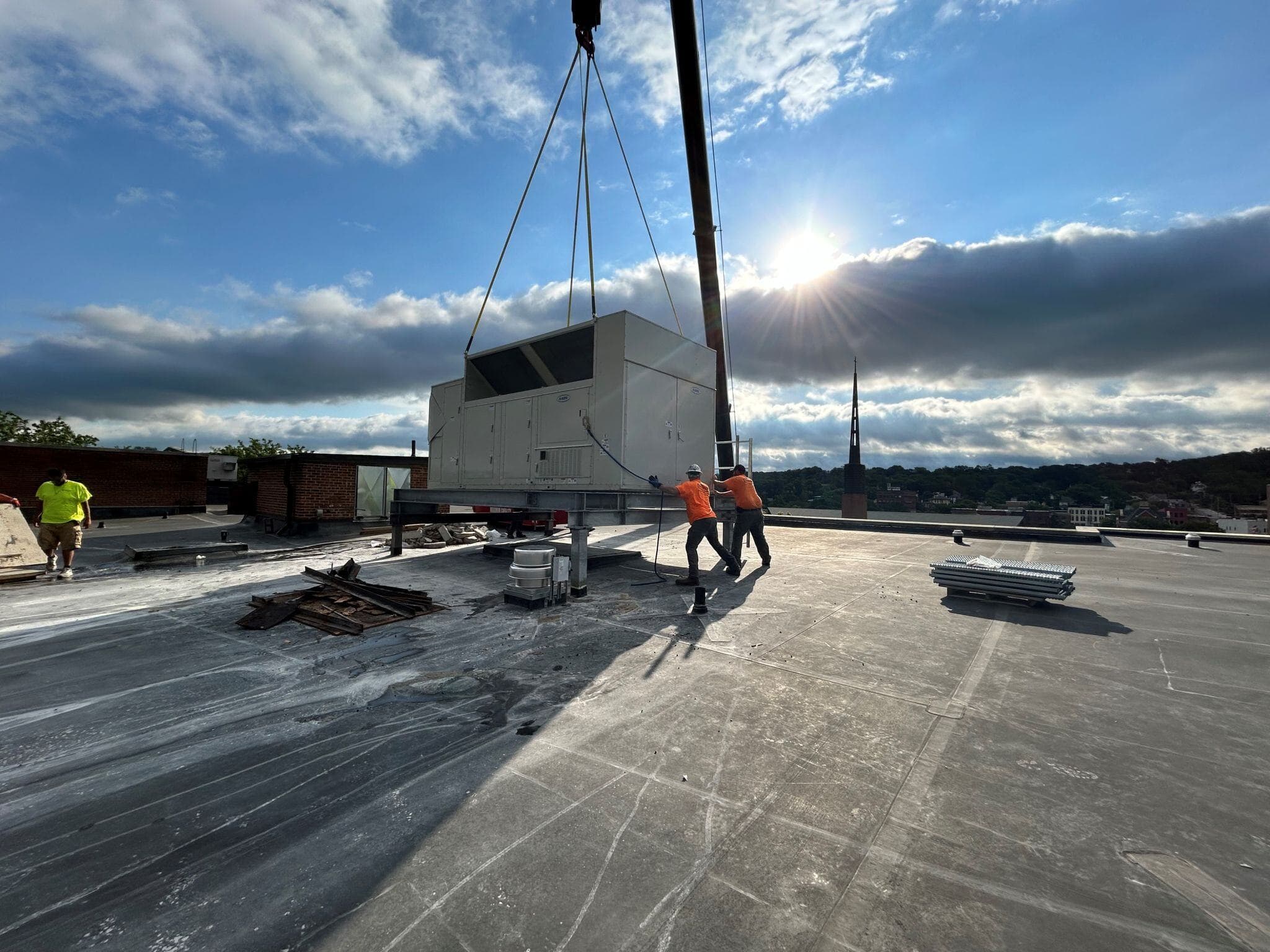 Construction crew guiding a rooftop HVAC unit into place with a crane on a commercial building under a bright sky.