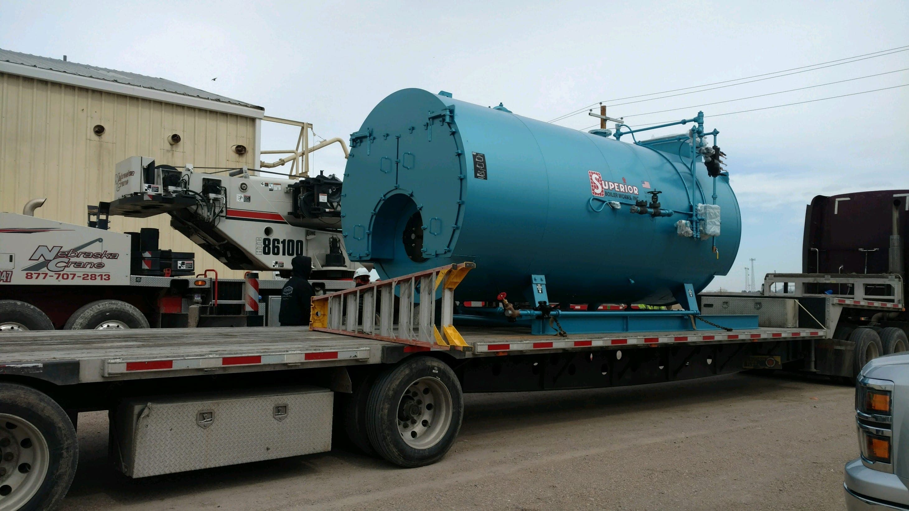 Large blue Superior industrial boiler loaded on a flatbed truck outside an industrial building.