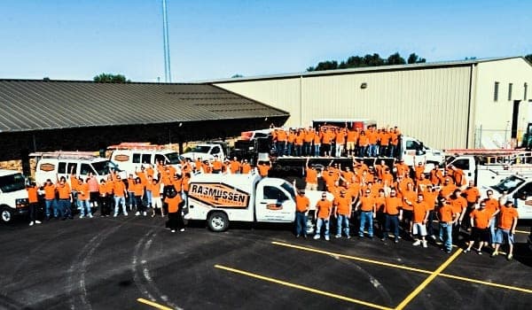 A large group of people in orange shirts stand in front of several white company vehicles in a parking lot near a beige industrial building.