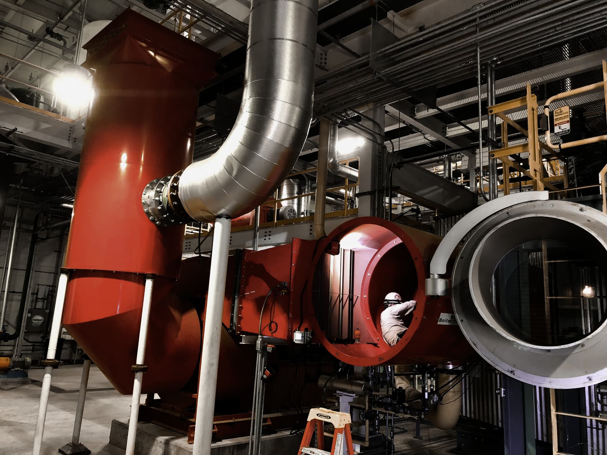 Technician working inside a large red industrial burner system with access panels open and ductwork above, inside a mechanical room with scaffolding and lighting.