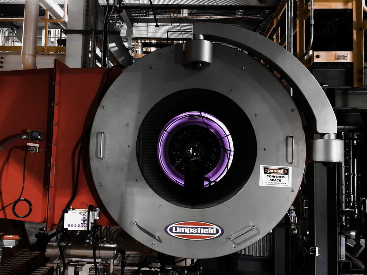Frontal view of a Limpsfield industrial burner with visible purple combustion flame, surrounded by pipes and control systems inside a mechanical room.